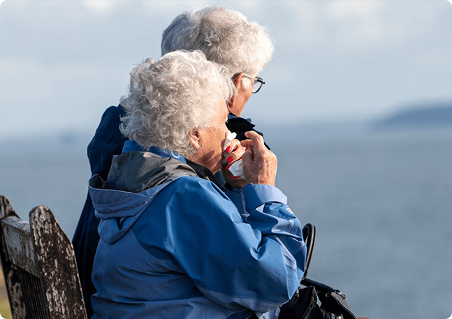 Two seniors on a bench by the ocean; one uses a tissue.
